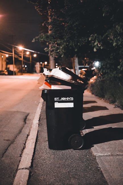 A sanitation worker in high-visibility orange trousers, a white jacket with reflective strips, and a blue cap is using a leaf blower with a black hose to clear household waste and litter from a grassy