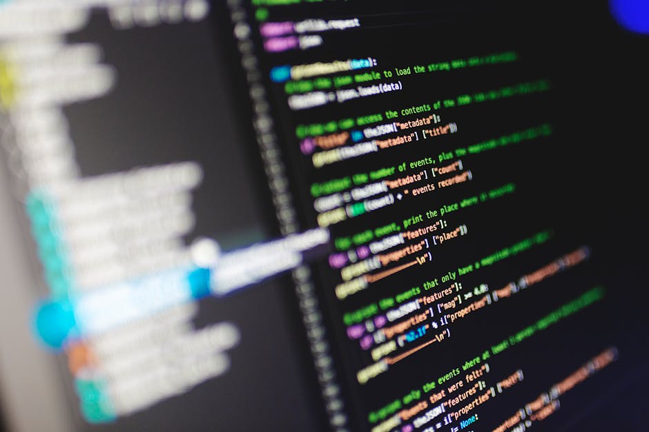 Close-up photograph of a computer screen displaying lines of colourful computer code text against a dark background, with various shades of green, purple, orange, and blue. The text appears organized 